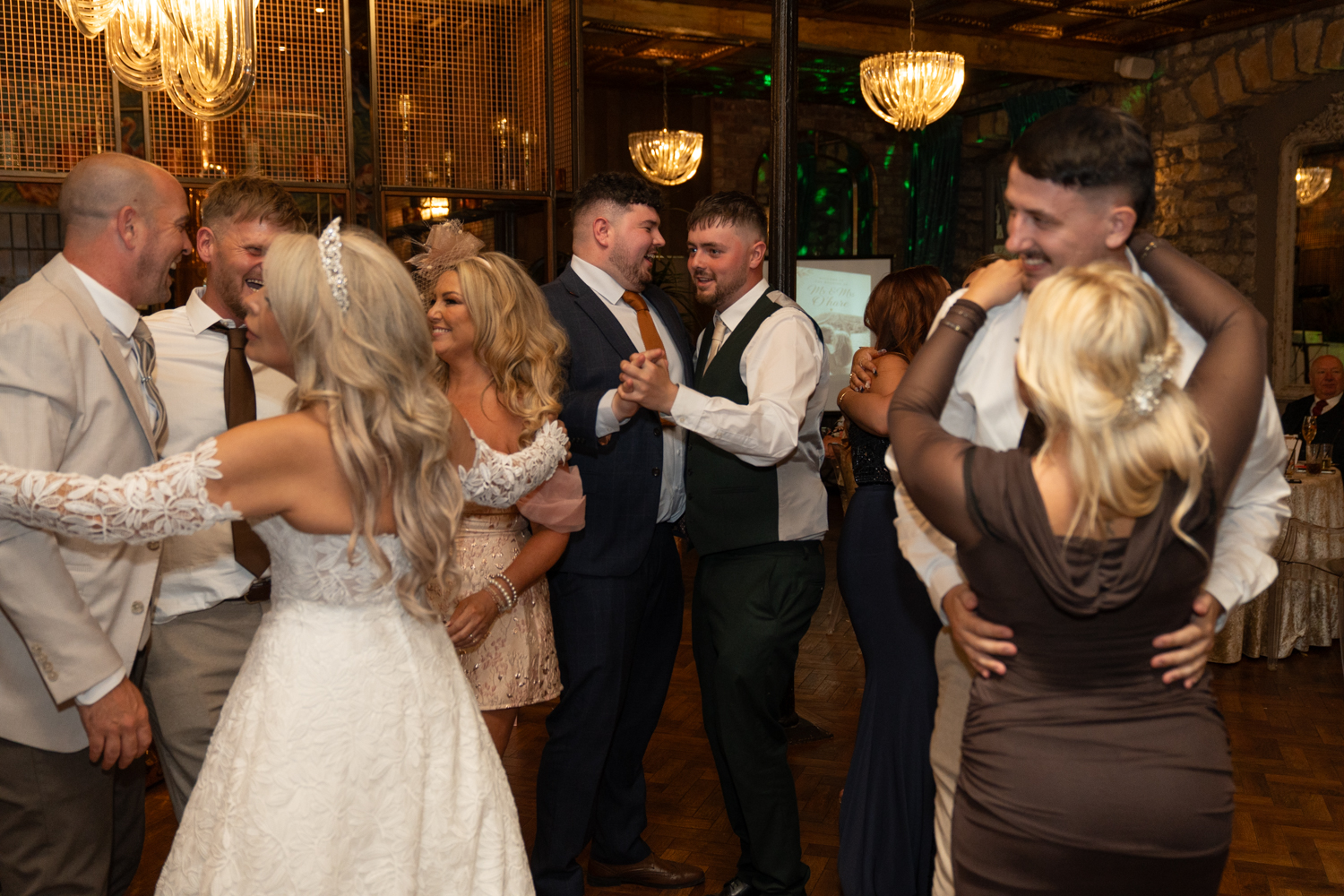The whole wedding party on the dance floor — bride and groom surrounded by family and friends, everyone laughing and moving together